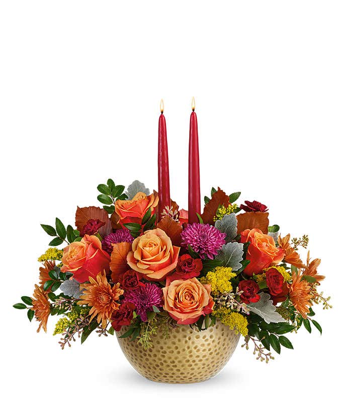 A rich floral arrangement featuring orange roses, maroon miniature carnations, rust and purple cushion spray chrysanthemums, solidago, brown copper beech, dusty miller, seeded eucalyptus, and huckleberry, complemented by burgundy candles, all beautifully 