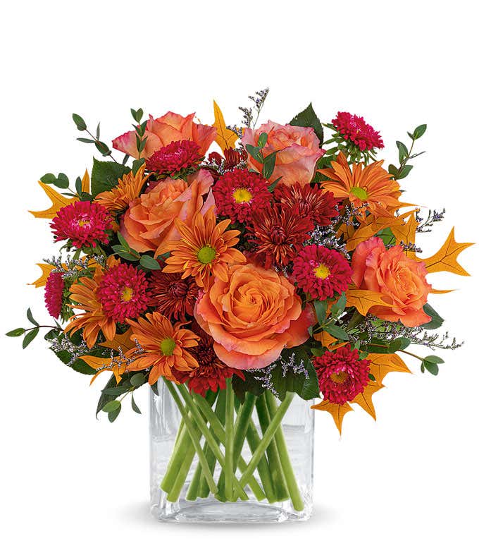 A vibrant floral arrangement featuring orange roses, red Matsumoto asters, bronze cushion spray chrysanthemums, bronze daisy spray chrysanthemums, purple limonium, parvifolia eucalyptus, and yellow oak leaves, all elegantly arranged in a bronze vase.