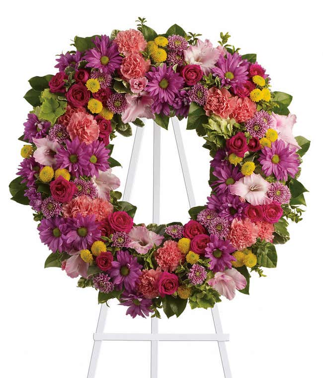 Funeral wreath with pink carnations, pink spray roses, green hydrangeas, pink gladioli, and green foliage on a white metal easel.