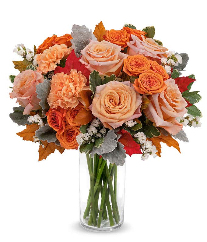 Peach and orange roses, orange carnations, white statice, variegated pittosporum, Red Oak Leaves in a brown ceramic pitcher against a white background