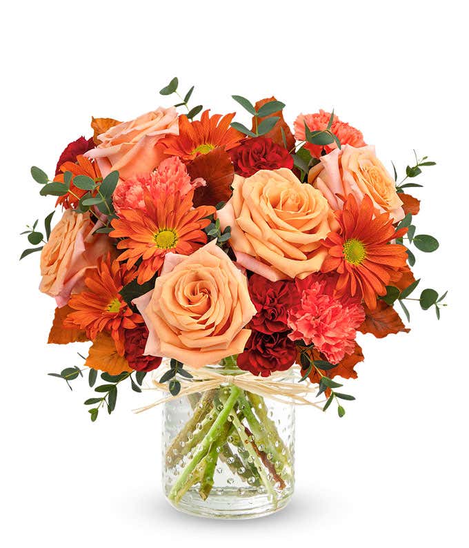 Light Orange Roses, Orange Carnations, Maroon mini carnations, bronze daisy spray chrysanthemums, brown copper beech, and parvifolia eucalyptus in a copper cylinder vase with patina against a white background