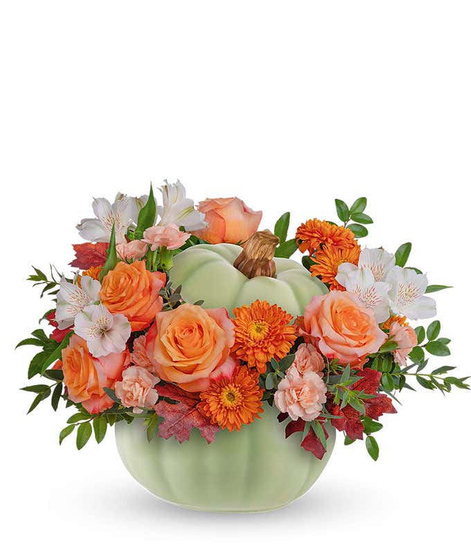 A fall floral arrangement featuring vibrant orange roses, white alstroemeria, miniature peach carnations, and bronze cushion spray mums, accented with seasonal greenery, all artfully arranged in a light green ceramic pumpkin-shaped container.