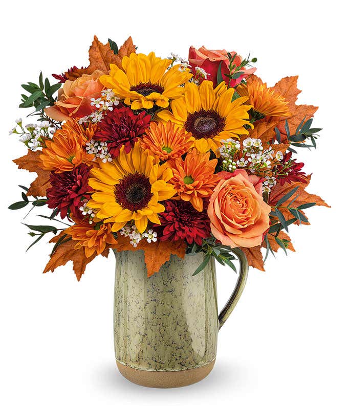 A cheerful floral arrangement featuring orange roses, red cushion spray mums, bronze daisy spray mums, and bright yellow sunflowers, accented with seasonal floral greenery, and presented in a rustic ceramic pitcher with a speckled finish.