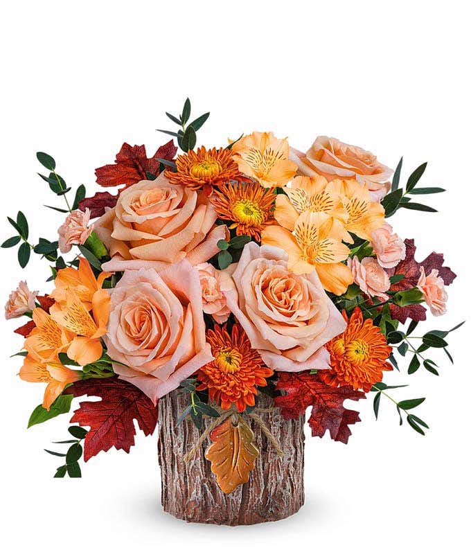 A rustic floral arrangement featuring peach roses, peach alstroemeria, peach miniature carnations, and bronze cushion spray mums, accented with seasonal floral greenery and presented in a bark-like vase with a decorative leaf accent.