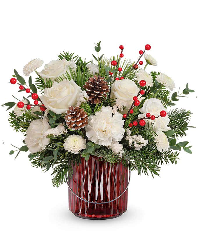 Holiday floral arrangement featuring white roses, white carnations, pinecones, red berries, and festive greenery, displayed in a red glass vase with silver accents, creating a wintery and elegant look.