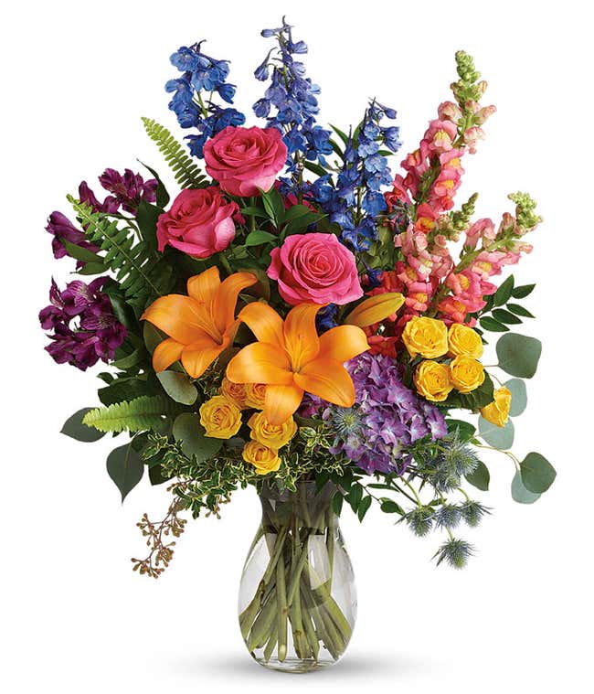 A vibrant arrangement featuring purple hydrangea, pink roses, yellow spray roses, orange Asiatic lilies, purple alstroemeria, blue delphinium, pink snapdragons, blue eryngium, huckleberry, eucalyptus, and fresh greenery, beautifully presented in a clear g