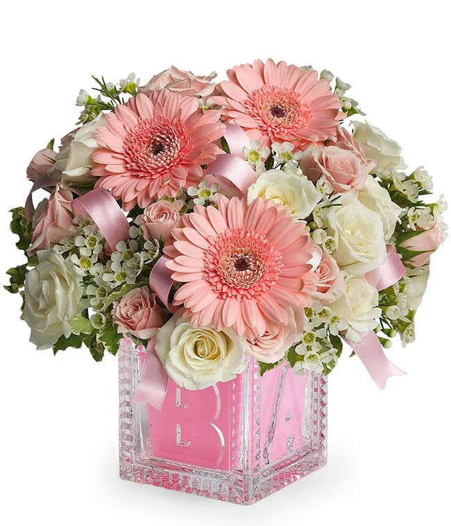 A floral arrangement featuring pink gerbera daisies, white spray roses, pink spray roses, and Monte Casino in a glass baby block vase.