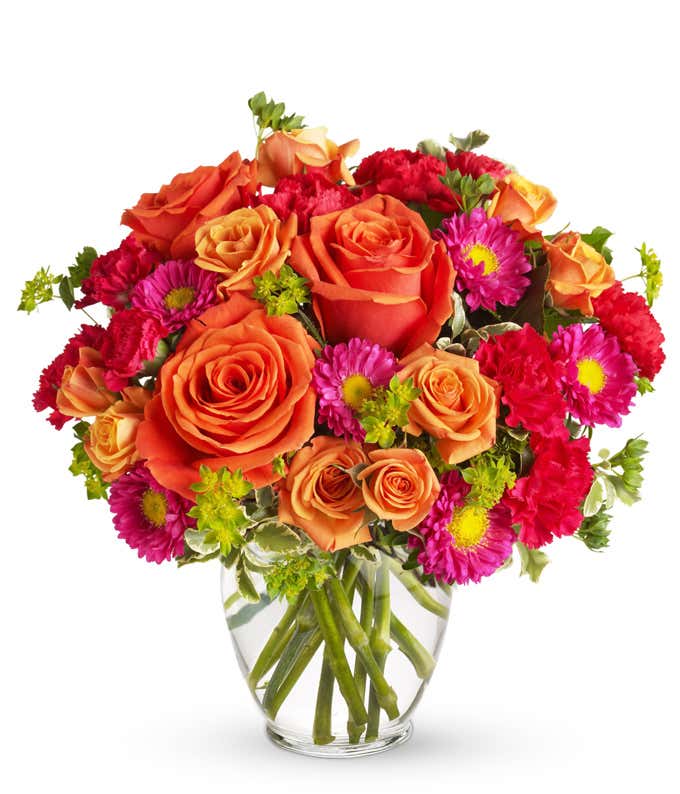 Floral arrangement featuring light orange roses, orange spray roses, hot pink carnations, and matsumoto asters in a clear glass vase.