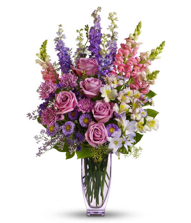 A beautiful lavender and pink arrangement in a lavender vase featuring roses, alstroemeria, snapdragons, larkspur, freesia, matsumoto asters, monte cassino asters, cushion spray mums, statice, and limonium. This elegant display offers a harmonious and soo