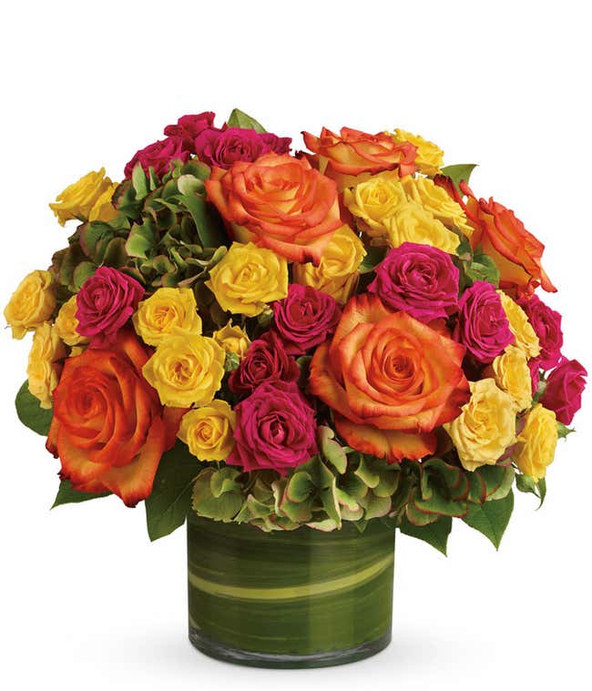 A floral arrangement featuring green hydrangea, hot pink roses, yellow roses, and bi-color roses in a clear glass vase.