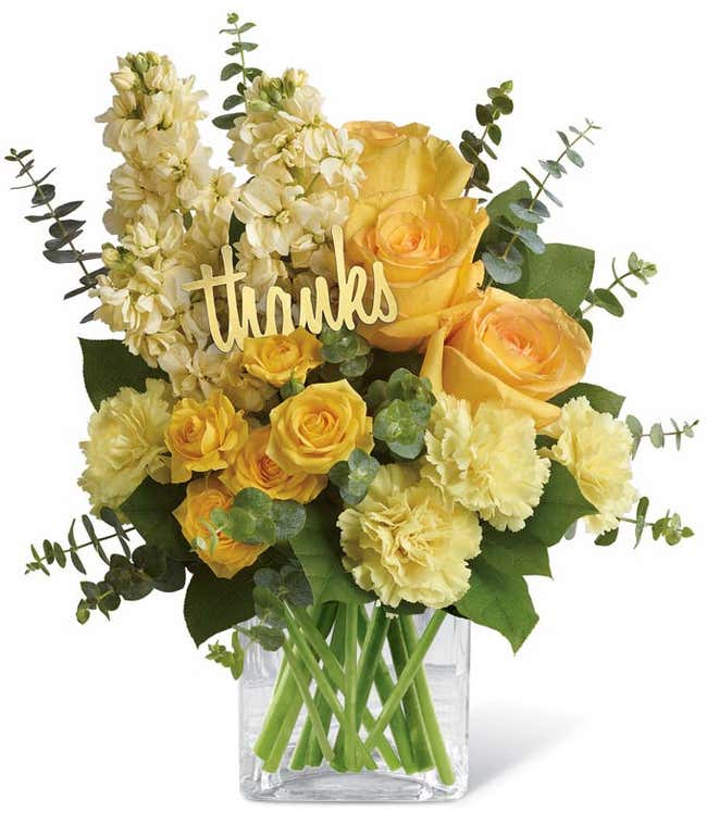 Floral arrangement featuring yellow roses, yellow spray roses, light yellow stock, spiral eucalyptus, lemon leaf, with a 'Thanks' pick, in a clear glass cube vase.