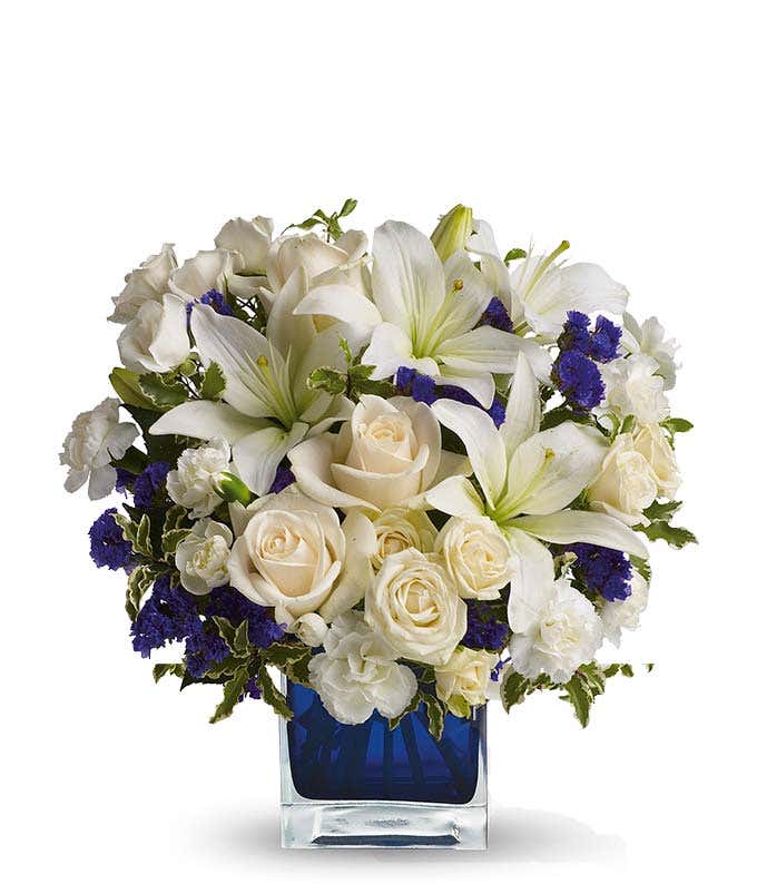 Floral arrangement featuring crme roses, crme spray roses, white Asiatic lilies, white miniature carnations, and purple statice in a blue cube vase.