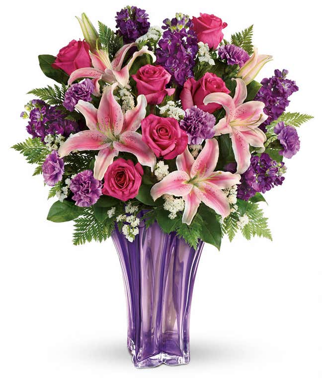 A floral arrangement featuring hot pink roses, pink oriental lilies, purple carnations, and floral greens in a keepsake lavender vase.