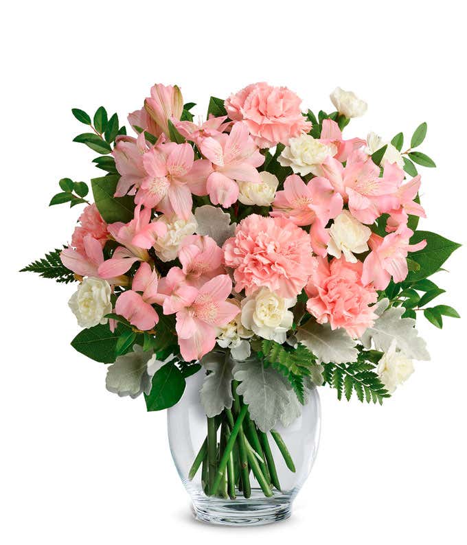 A delicate floral arrangement of pink alstroemeria, pink carnations, white mini carnations, dusty miller, huckleberry, leatherleaf fern, and lemon leaf, elegantly arranged in a clear glass vase. This soft blend offers a gentle and inviting display.