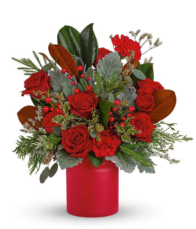 A festive arrangement with red roses, red carnations, red spray roses, red berries, white limonium, seasonal greens, displayed in a red vase.