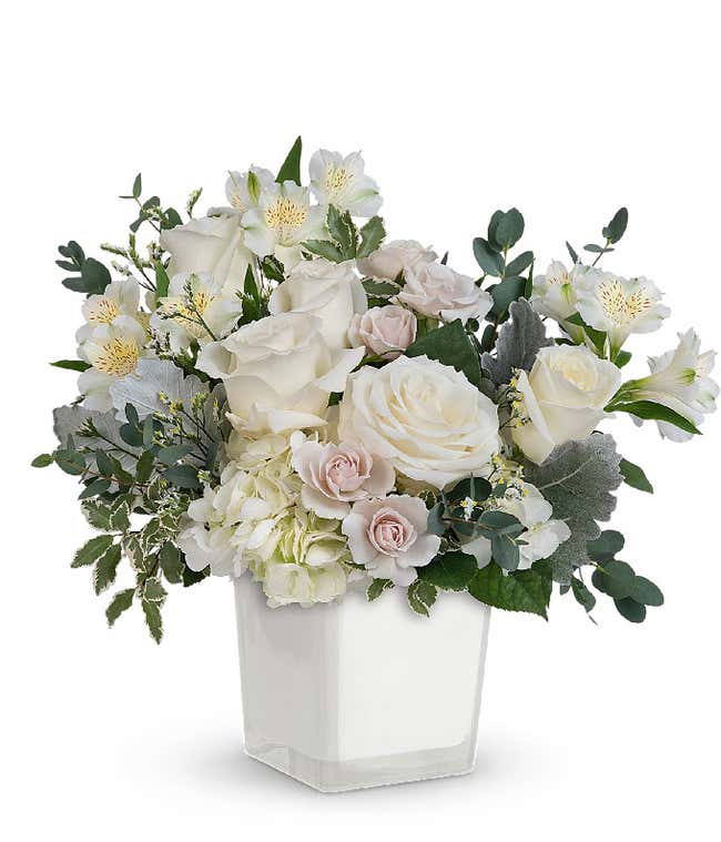 A serene arrangement featuring white hydrangea, white roses, white spray roses, white alstroemeria, white limonium, and floral greens, all elegantly presented in a white cube. This all-white display offers a timeless and peaceful touch to any occasion.