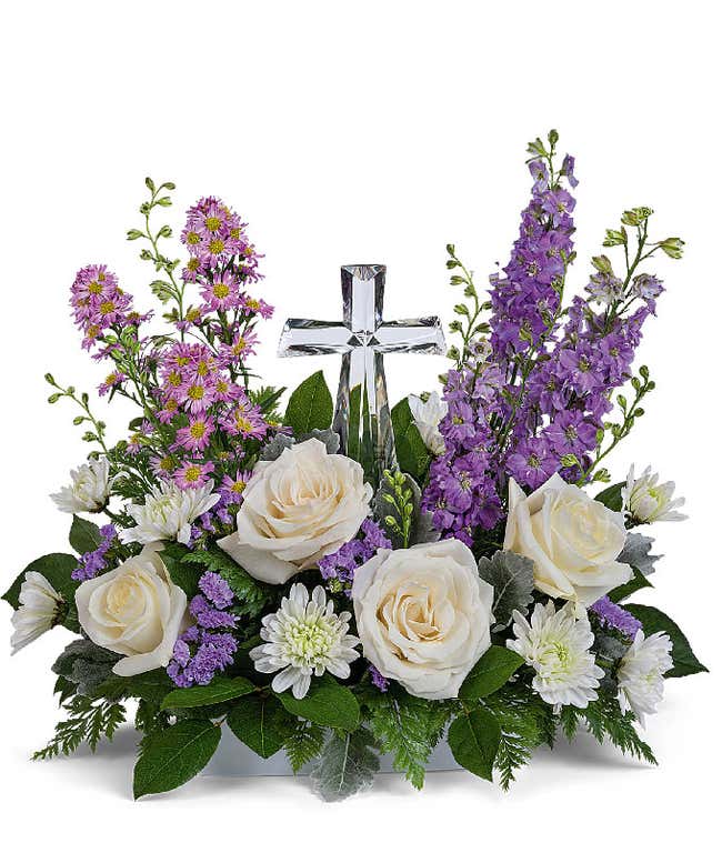 A graceful floral arrangement showcasing cr&egrave;me roses, lavender larkspur, lavender asters, lavender statice, white mums, and floral greens, elegantly accented with a crystal cross. This bouquet blends soft, calming colors for a serene display.