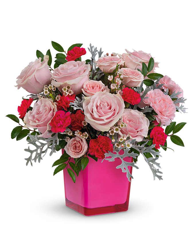 Pink roses, carnations, hot pink carnations, white waxflower, silver lace dusty miller, and huckleberry in a pink glass cube vase against a white background