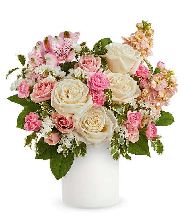 Delicate floral arrangement with ivory roses, light pink spray roses, pink alstroemeria, and pale peach stock in a sleek white vase, perfect for expressing love and care.