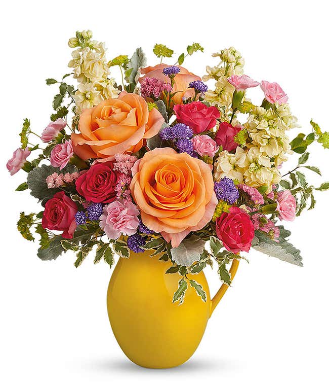 A cheerful bouquet of peach roses, hot pink spray roses, miniature pink carnations, yellow stock, and purple monte cassino, arranged in a keepsake yellow pitcher. A perfect gift for any occasion.