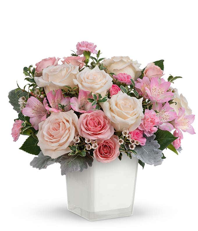 A floral arrangement featuring crme roses, pink spray roses, pink alstroemeria, pink miniature carnations, white waxflower, and greenery in a white vase.