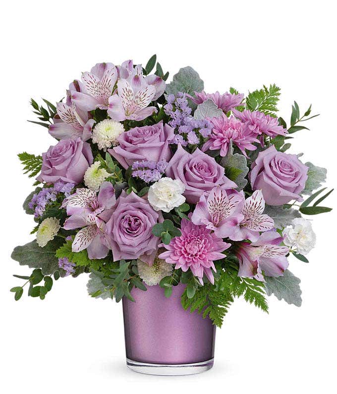 A floral arrangement of lavender roses, alstroemeria, miniature white carnations, white button spray chrysanthemums, lavender cushion spray chrysanthemums, and greenery in a purple cylinder container.