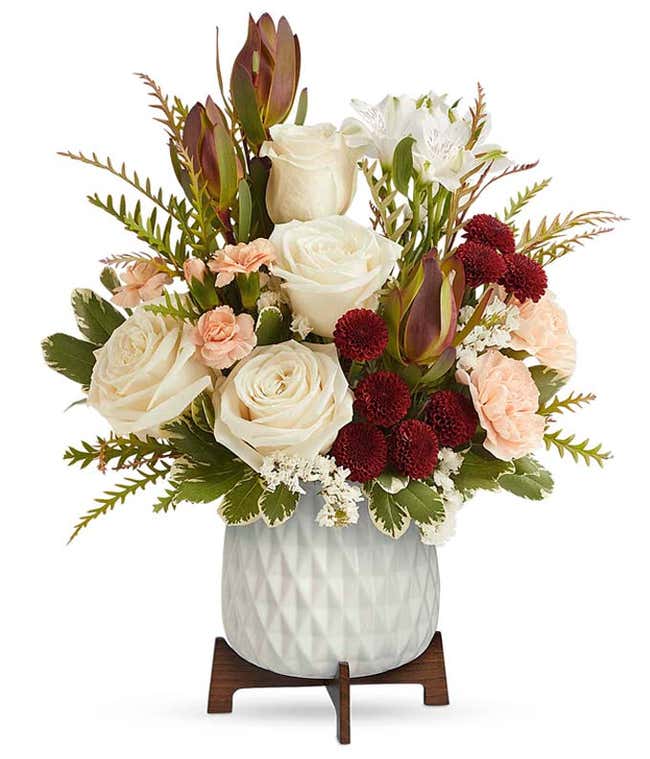 A bouquet of cr&egrave;me roses, white alstroemeria, red leucadendron, peach carnations, and burgundy button spray chrysanthemums in a white vase with a wooden stand.