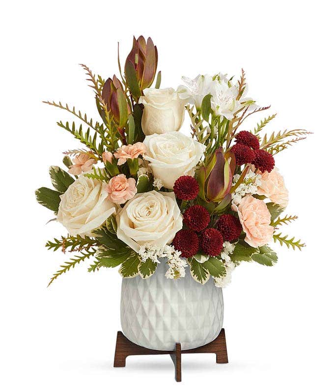 A bouquet of crme roses, white alstroemeria, red leucadendron, peach carnations, and burgundy button spray chrysanthemums in a white vase with a wooden stand.