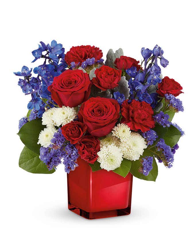 Patriotic floral arrangement with red roses, red carnations, blue delphiniums, blue statice, and white chrysanthemums in a sleek red glass vase, perfect gift for celebrating national pride.