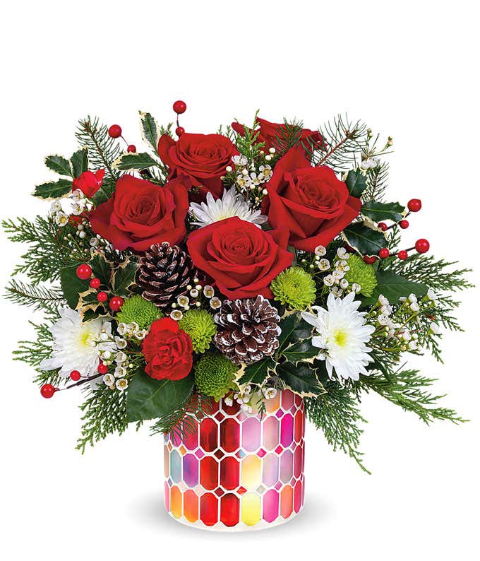 A festive arrangement with red roses, red miniature carnations, white cushion spray chrysanthemums, green button spray chrysanthemums, white waxflower, floral greens, displayed in a keepsake red mosaic cylinder vase.