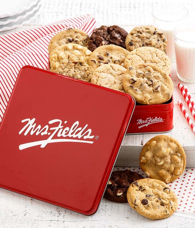  Red Mrs. Fields tin filled with a variety of cookies, including chocolate chip, white chocolate macadamia, and double chocolate, displayed with glasses of milk and striped straws on a white wooden surface.