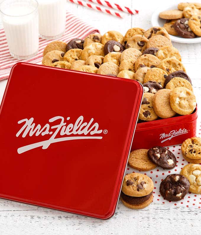  Red Mrs. Fields tin filled with an assortment of mini cookies, including chocolate chip, white chocolate macadamia, and double chocolate, arranged on a white wooden surface with glasses of milk and striped straws in the background.