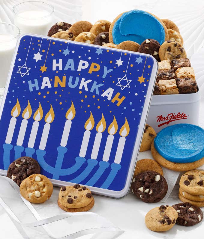 Hanukkah-themed cookie gift tin featuring a blue design with a menorah, candles, and Happy Hanukkah text surrounded by stars and dots. The tin is filled with assorted cookies, including chocolate chip, brownie bites, and frosted blue sugar cookies.