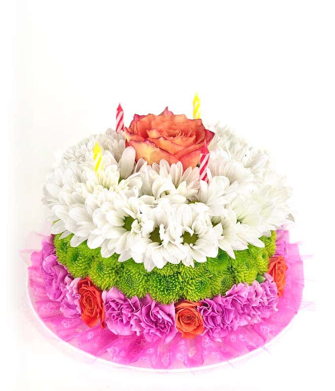 A floral arrangement designed to resemble a birthday cake, featuring layers of white daisies, green button mums, and pink carnations. The top layer is accented with a single peach rose and three striped candles, with a decorative pink lace trim around the