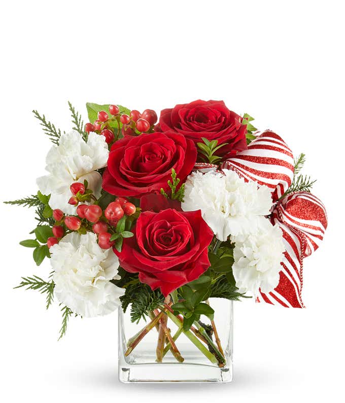 A festive arrangement with red roses, white carnations, red hypericum berries, displayed in a glass cube vase with a decorative ribbon.