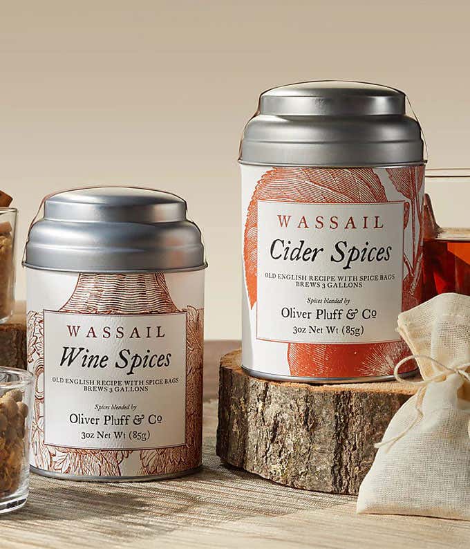 Wassail gift set featuring two tins: Wine Spices and Cider Spices, displayed on a rustic wood slice. The tins are designed for brewing traditional wassail and include spice bags. The scene is complemented by dried fruits and a tied muslin bag.